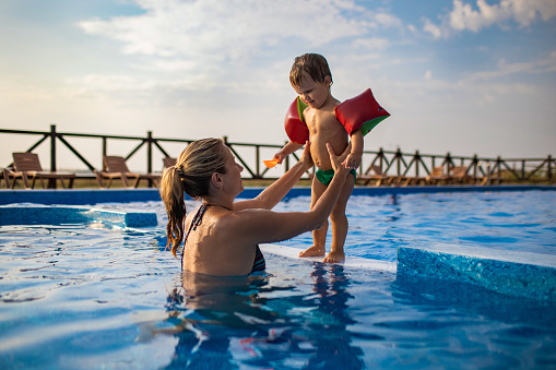 Beautiful happy cheerful mom plays with her little funny cute son with bright colorful oversleeves in a deep blue pool with clear transparent water, in summer
