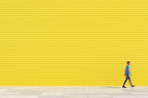 Man in blue clothes walks on the sidewalk in front of a blank yellow colour wall, Copy space background. 3D - Computer generated image.
