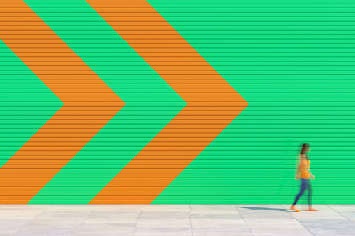 A woman in an orange dress walks on the sidewalk in front of a colorful wall in the direction of the arrow, the person is blurred, Copy space background. 3D - computer-generated image.