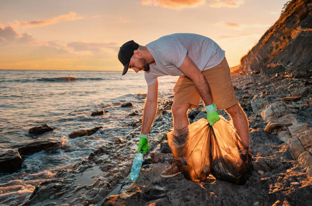 Cleaning beach from the pollution. Volunteer picking up a plastic bottle. In the background, the sea and the sunset. Copy space. The concept of environmental conservation Cleaning beach from the pollution. Volunteer picking up a plastic bottle. In the background, the sea and the sunset. Copy space. The concept of environmental conservation. earth day clean up stock pictures, royalty-free photos & images