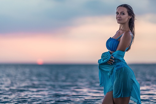 A slender tanned girl with wet tousled hair in a gentle blue swimsuit and a bright pareo, poses against the background of a warm sunset in a clean estuary with transparent water