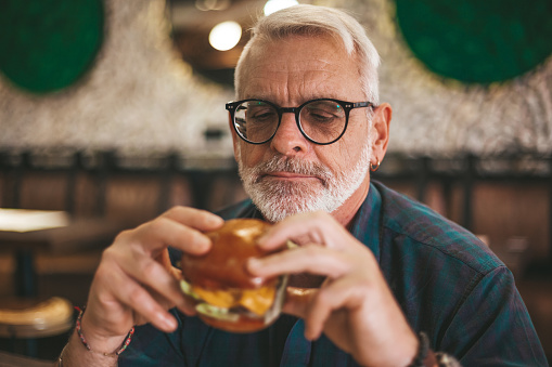 Senior gray-haired man in glasses with a beard is eating an appetizing burger. Lunch break, rest in the pub.