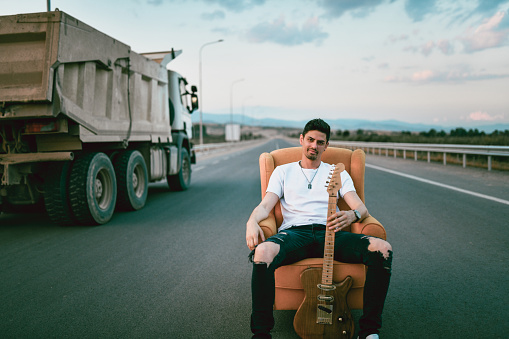 Guitarist Sitting On Chair With Guitar On Highway While Truck Passes By