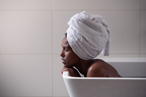 Portrait of a beautiful Afro-American woman with a white towel on her head relaxing in a bathtub. Female taking bath in the bathroom.