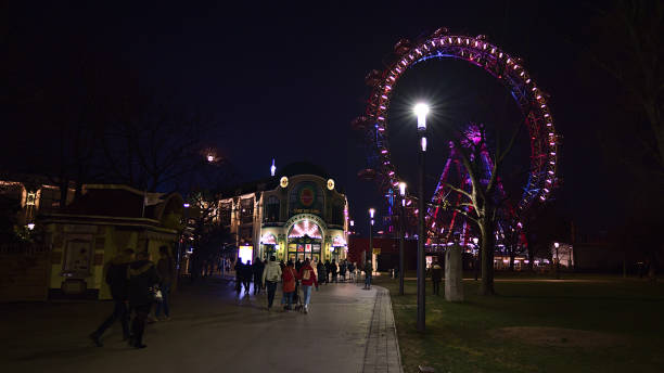 vue nocturne des gens marchant jusqu’à l’entrée du parc d’attractions wurstelprater à vienne, en autriche, avec la célèbre grande roue éclairée (riesenrad) et un lampadaire lumineux. - vienna ferris wheel night prater park photos et images de collection