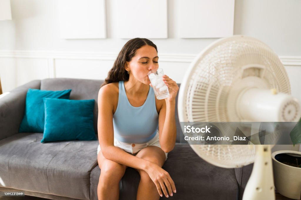 Woman enjoying a cold drink in front of the fan Happy woman trying to cool down drinking an ice cold drink and turning on the electric fan at home Heat Wave Stock Photo