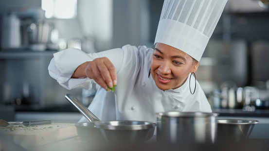 Restaurant Kitchen: Black Female Chef Fries Uses Pan, Seasons Dish with Herbs and Spices, Smiles. Professional Cooking Delicious and Traditional Authentic Food. Healthy Dishes Prepare. Medium Shot