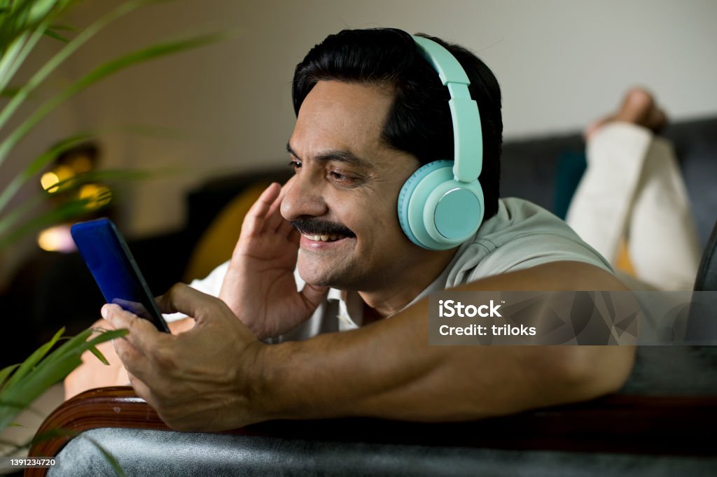 Man listening music or watching video using mobile phone and headphones Happy man listening music and watching video on mobile phone while lying down on sofa with headphones Adult Stock Photo
