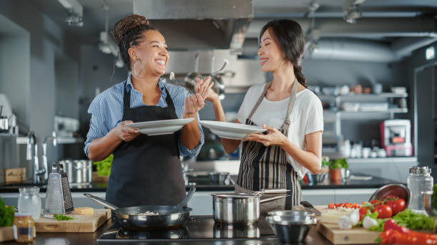 tv cooking show kitchen with two master chefs. asian and black female hosts talk. professionals teach how to cook food, taste delicious dish. online video class courses. healthy dish recipe prepare - playback imagens e fotografias de stock