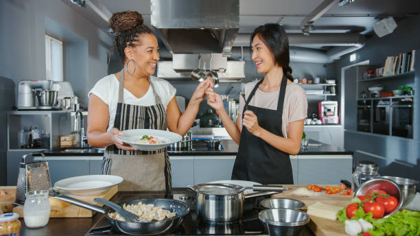 tv cooking show kitchen with two master chefs. asian and black female presenters talk on camera, teach how to cook food, taste delicious dish and celebrate with high-five. online video class - chef’s utensils imagens e fotografias de stock