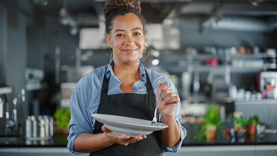 Restaurant Kitchen: Portrait of Black Female Chef Preparing Dish, Tasting Food and Enjoying it. Professional Cooking Delicious, Authentic, traditional Food, using Healthy Ingredients for Meal Recipe