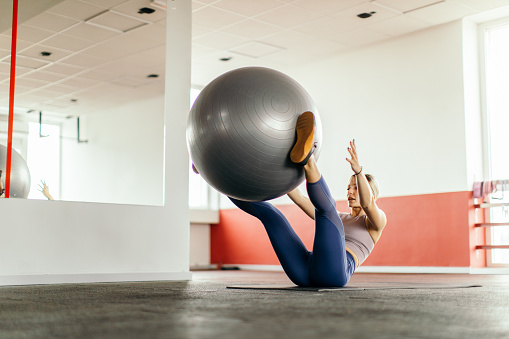 Young blonde woman exercising with fitness ball and looks very concentrated and motivated. She wears sports clothes and exercising in gym and she looks as real woman who likes to live healthy lifestyle.