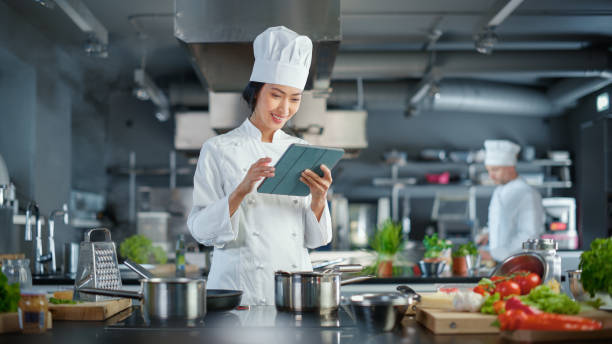 ristorante di fama mondiale: chef asiatico che cucina cibo delizioso e autentico, utilizza un tablet digitale mentre lavora in una moderna cucina professionale. preparazione di piatti biologici gourmet - cucina commerciale foto e immagini stock