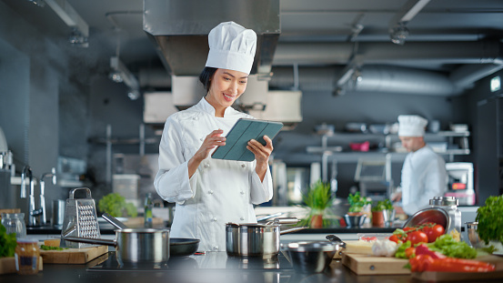 World Famous Restaurant: Asian Female Chef Cooking Delicious and Authentic Food, Uses Digital Tablet Computer While Working in a Modern Professional Kitchen. Preparing gourmet organic Dishes