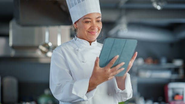 レストランキッチン:おいしいと本物の食品を調理黒人女性シェフ、デジタルタブレットコンピュータを使用しています。プロフェッショナルは、健康的な食材を使用して、伝統的な料理を作 - 女性料理人 ストックフォトと画像