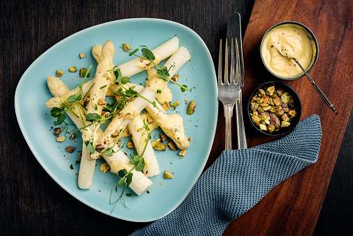 Overhead view of a plate of white asparagus with a pistachio and hollandaise sauce served with fresh pea shoots. The white asparagus has been peeled and the last few woody centimetres of the stalk removed and then cooked gently in a mix of boiling water, lemon juice and salt for between 8-12 minutes depending on the thickness of the stalk. Here is a served with fresh hollandaise sauce, fresh pea shoots and some chopped pistachio nuts. The pistachios give a nice crunch to contrast with the softness of the the asparagus. Colour, horizontal format with some copy space.