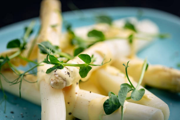 Close-up view of a plate of white asparagus with a pistachio and hollandaise sauce served with fresh pea shoots. The white asparagus has been peeled and the last few woody centimetres of the stalk removed and then cooked gently in a mix of boiling water, lemon juice and salt for between 8-12 minutes depending on the thickness of the stalk. Here is a served with fresh hollandaise sauce and fresh pea shoots. Colour, horizontal format with some copy space.