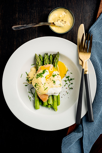 Overhead view of a plate of asparagus served with smoked salmon, poached egg with a hollandaise sauce. The last few woody centimetres of the asparagus stalks have been removed and they have been cooked gently in a mix of boiling water, lemon juice, and salt for between 8-12 minutes depending on the thickness of the stalk. A poached egg is placed on top and served with fresh hollandaise sauce and fresh chives. Colour, vertical format with some copy space.