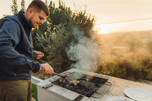 One young man enjoying the beautiful sunset over tree area from his house terrace preparing grilled meat on fire for picnic