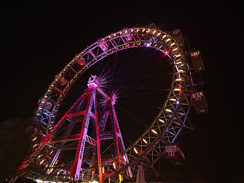 Stunning low angle view of popular Ferris wheel with colorful illuminated rim at night in amusement park Prater (Wurstelprater) in Vienna, Austria. Viewed from public ground.