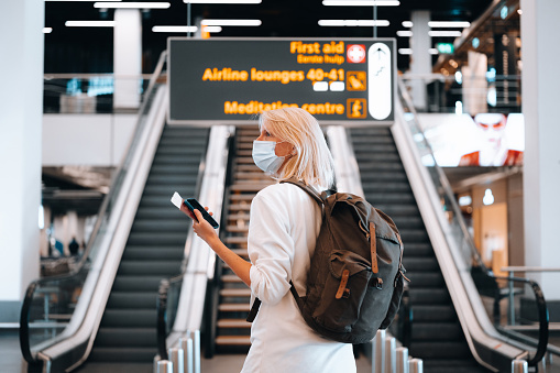 Rear view of a woman at the airport near the escalator, holding a passport with a boarding pass and a mobile phone