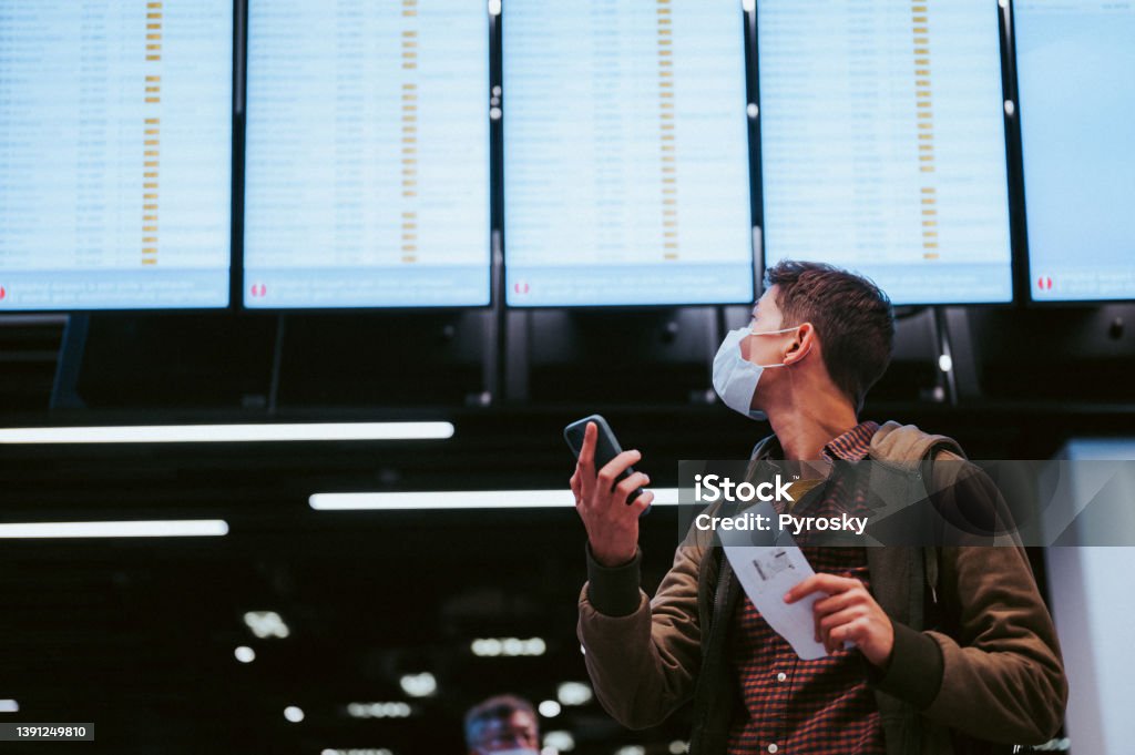 Checking the flight schedule A low angle view of a young man checking his flight on the departure board.
He holds a mobile phone and a plane ticket in his hands. Travel concept Nomadic People Stock Photo