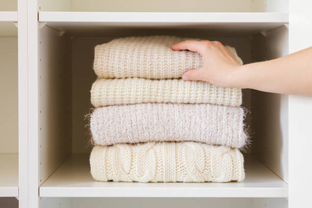Young adult woman hand taking folded knitted light sweater from shelf in white opened wardrobe at room. Front view. Closeup. Female wool warm clothes. stock photo