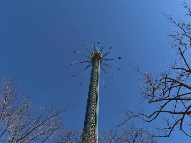 Photo of Beautiful low angle view of rotating drop tower in popular amusement park Wurstelprater in Vienna, Austria with bare trees in front on sunny day in spring.