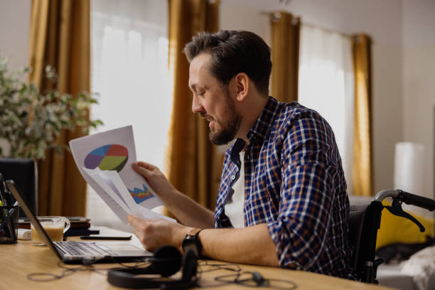 un adulto brunet su una sedia a rotelle sfoglia i documenti aziendali, sfogliando ogni pagina. un laptop si trova di fronte a lui. l'uomo è sorridente e felice del suo lavoro a casa - twitter business web page browser foto e immagini stock