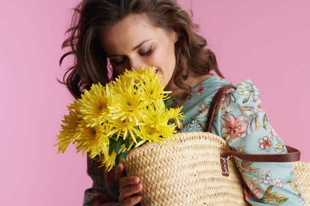 smiling trendy woman in floral dress on pink smiling trendy middle aged woman in floral dress with yellow chrysanthemums flowers and straw bag against pink background. straw bag stock pictures, royalty-free photos & images