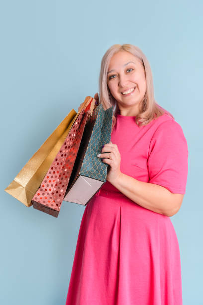 dorosła blondynka w wieku 40 lat trzyma w rękach wiele toreb z zakupami stojąc na niebieskim tle, pionowym zdjęciu - 35 40 years women female smiling zdjęcia i obrazy z banku zdjęć
