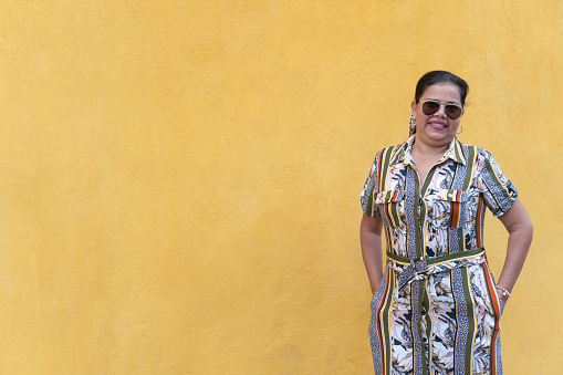 Latin woman in a dress and sunglasses stands against a yellow wall