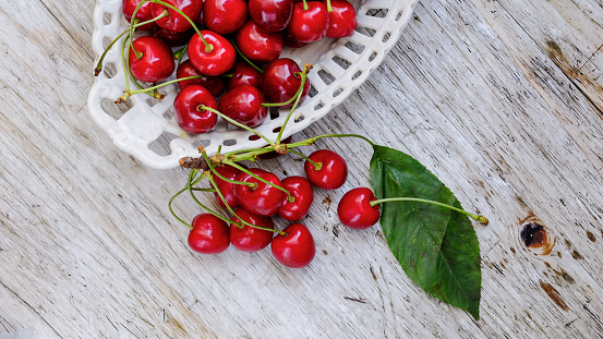 Cherries with cherry tree leaves placed in a porcelain bowl on a towel on a wooden table, close-up