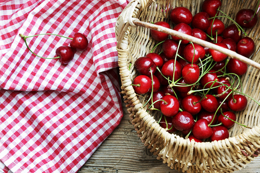 Cherries with cherry tree leaves in a basket placed on a towel on a wooden table, close-up gray