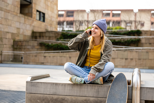 Young blonde woman with veri peri coolor hat talking with friend using smart phone, teenager female skater sitting on a bench in the city, copy space