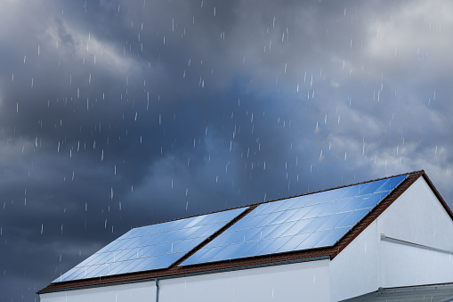 Top part of a building covered with large solar panels. Overcast sky with heavy clouds and rain.