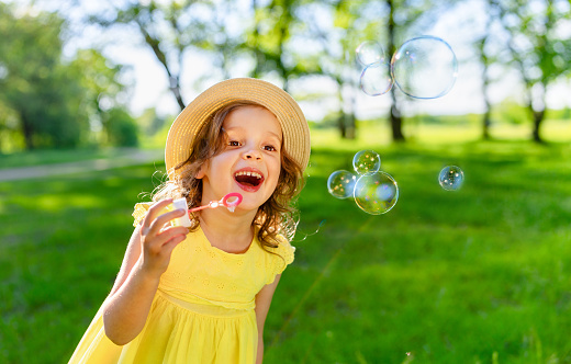 a carefree laughing girl in a bright yellow dress and a panama hat blows soap bubbles on a bright sunny day.