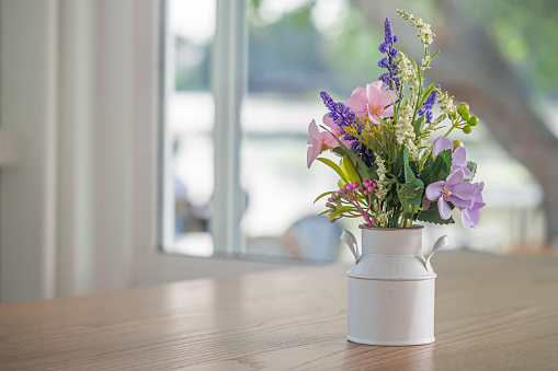 Closeup of white vintage vase of pot with colorful flower on wooden table and blurry window, tree and bokeh light