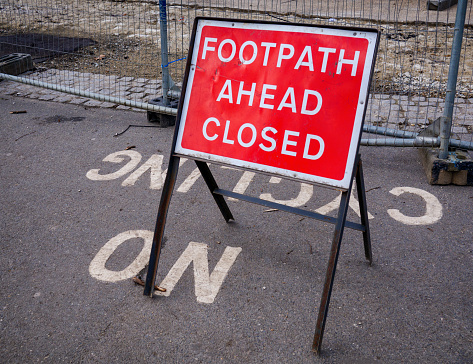 A red and white sign reading ‘Footpath Ahead Closed’ standing on tarmac with ‘No Cycling’ inscribed upon it.