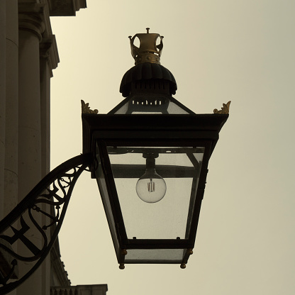 An old-fashioned lamp with a modern light bulb seen in silhouette against the sky. The lamp has a crown upon the top and an ornamental bracket attached to a stone column.