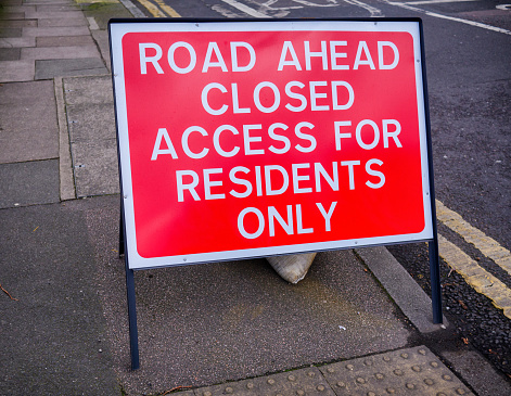 A red and white sign reading ‘Road Ahead Closed - Access for Residents Only’ standing on a pavement beside a road.