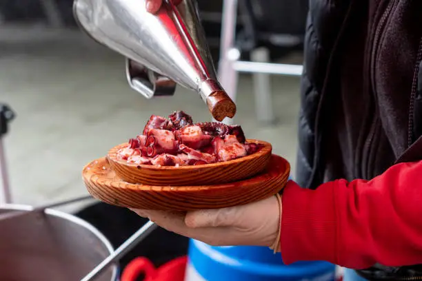 Photo of A man's hand pouring oil over a portion of octopus, prepared in the traditional Pulpo a feira style. Galicia, Spain.