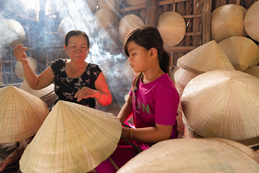 old vietnamese woman making with her grandchild a traditional conical hat at her home
