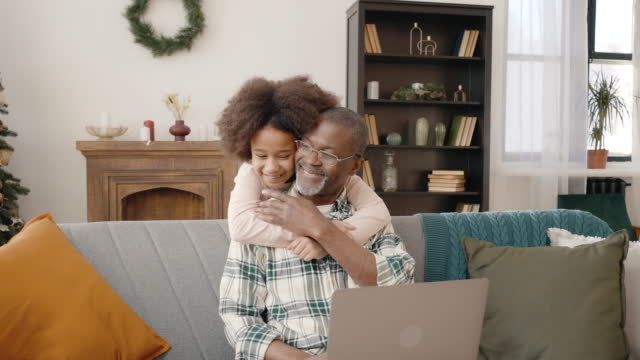 Focused senior african american man working on laptop at home, happy curly girl running and embracing her grandfather