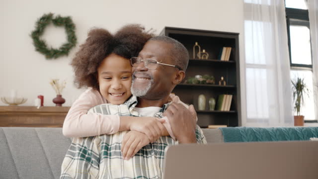 Lovely african american girl coming and embracing her busy grandfather, man in eyeglasses working on laptop at home