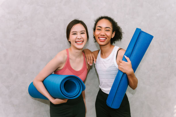 jovens mulheres adultas carregando tapete de yoga, sorrindo e emoção feliz, exercitando-se em casa com amigos , conceito de esporte e recreação. - friendship women exercising gym - fotografias e filmes do acervo