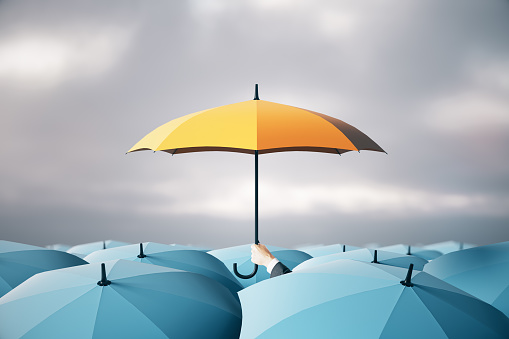 Businessman hand holding yellow umbrella over crowd on blurry dull sky background. Risk, protection and safety concept