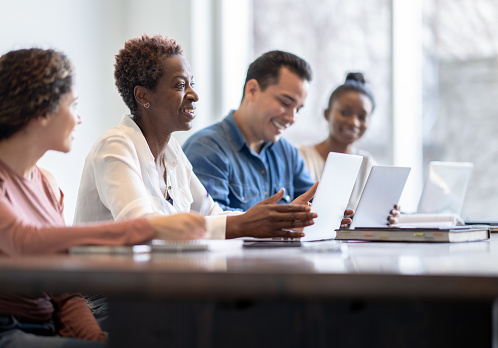 A small group of four mature adult students sit together at a long desk as they listen attentively to the teacher. They have text books and laptops out in front of them as they take notes and patriciate in the class and each is dressed casually.