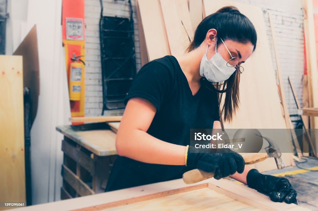 young woman working young woman wearing gloves and using a hammer, working in a carpentry shop, wearing a covid mask. Colombia Stock Photo