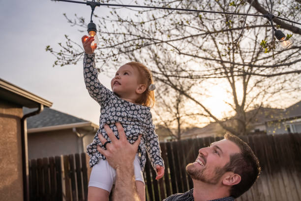 Young Father Holds Up Two-year Old daughter as she touches an edison light bulb decoratively arranged in a back yard at dusk stock photo
